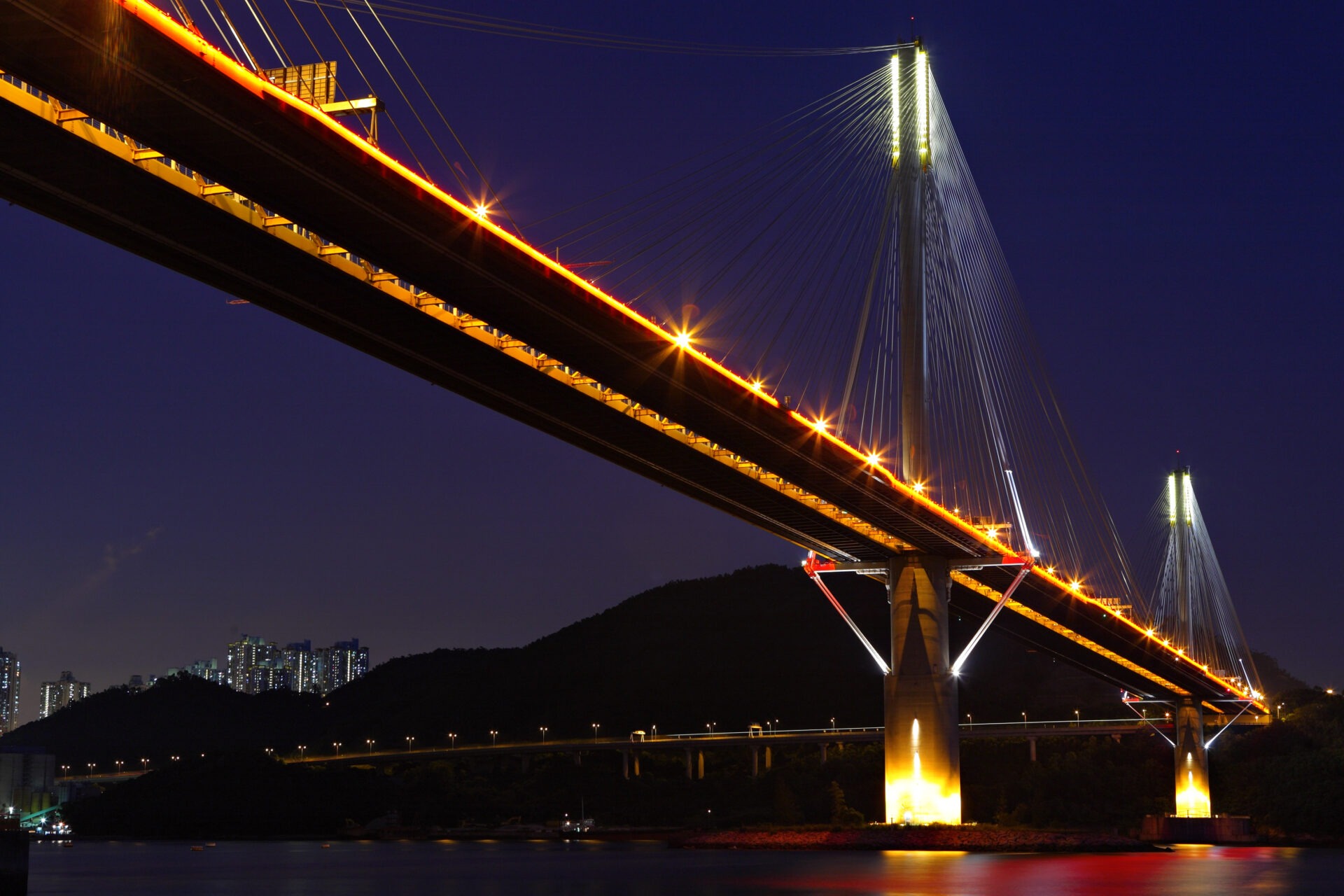 A cable-stayed bridge at night, lit along its cables, with foundation piers rising from dark water.